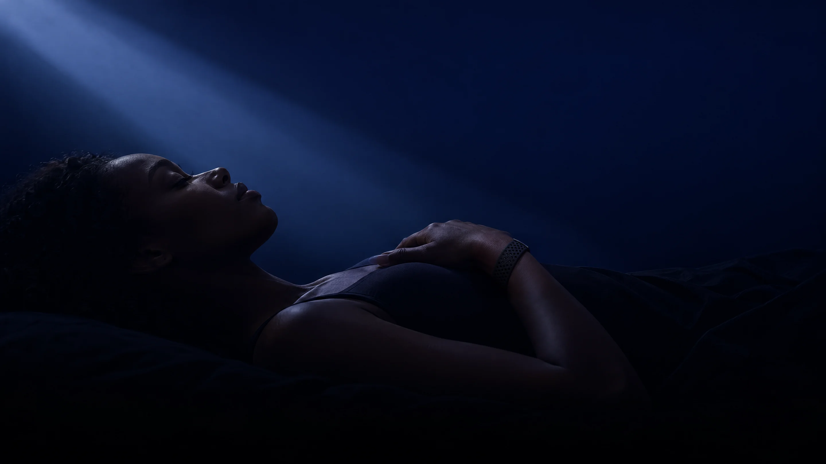A woman asleep on her back in a beam of moonlight, one hand resting on her chest, wearing the PhenomeTech Band in Onyx