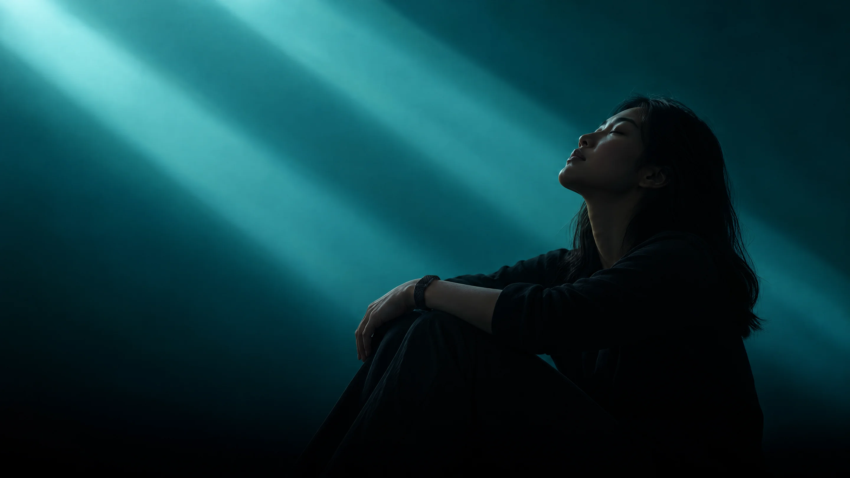 A woman seated with her knees drawn up, head tilted back and eyes closed in beams of teal light, wearing the PhenomeTech Band in Onyx