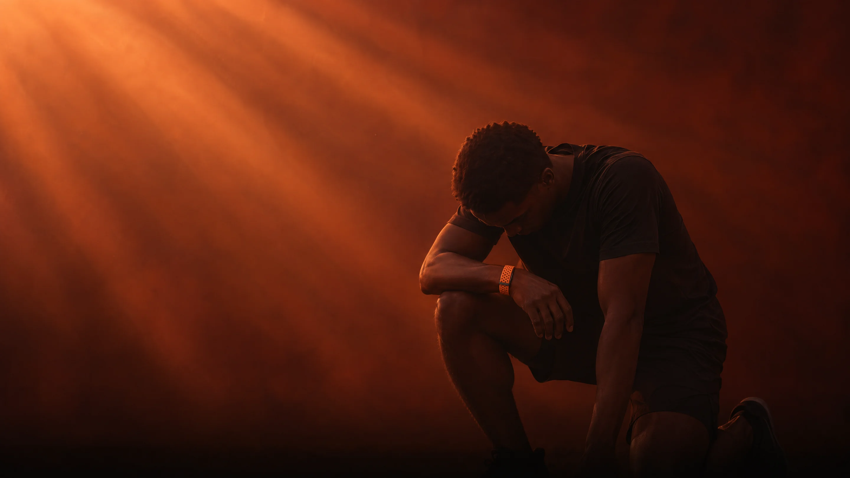 A man resting on one knee with his head lowered, catching his breath in soft beams of ember light, wearing the PhenomeTech Band in Ember
