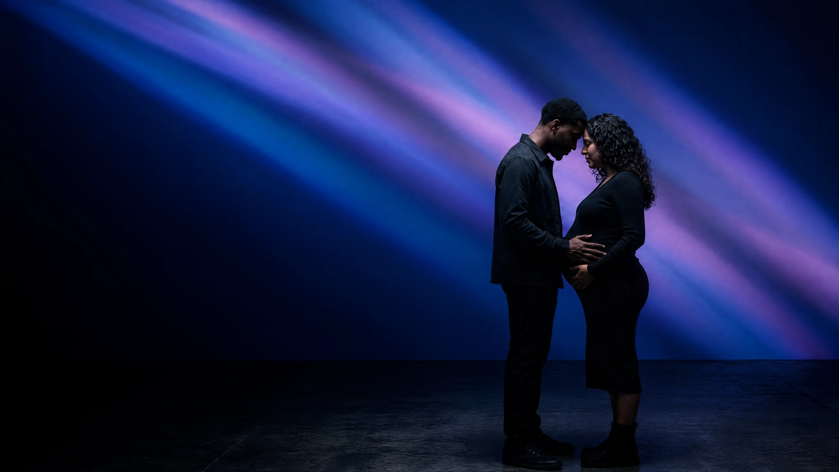 An expecting couple with their foreheads together in beams of violet light