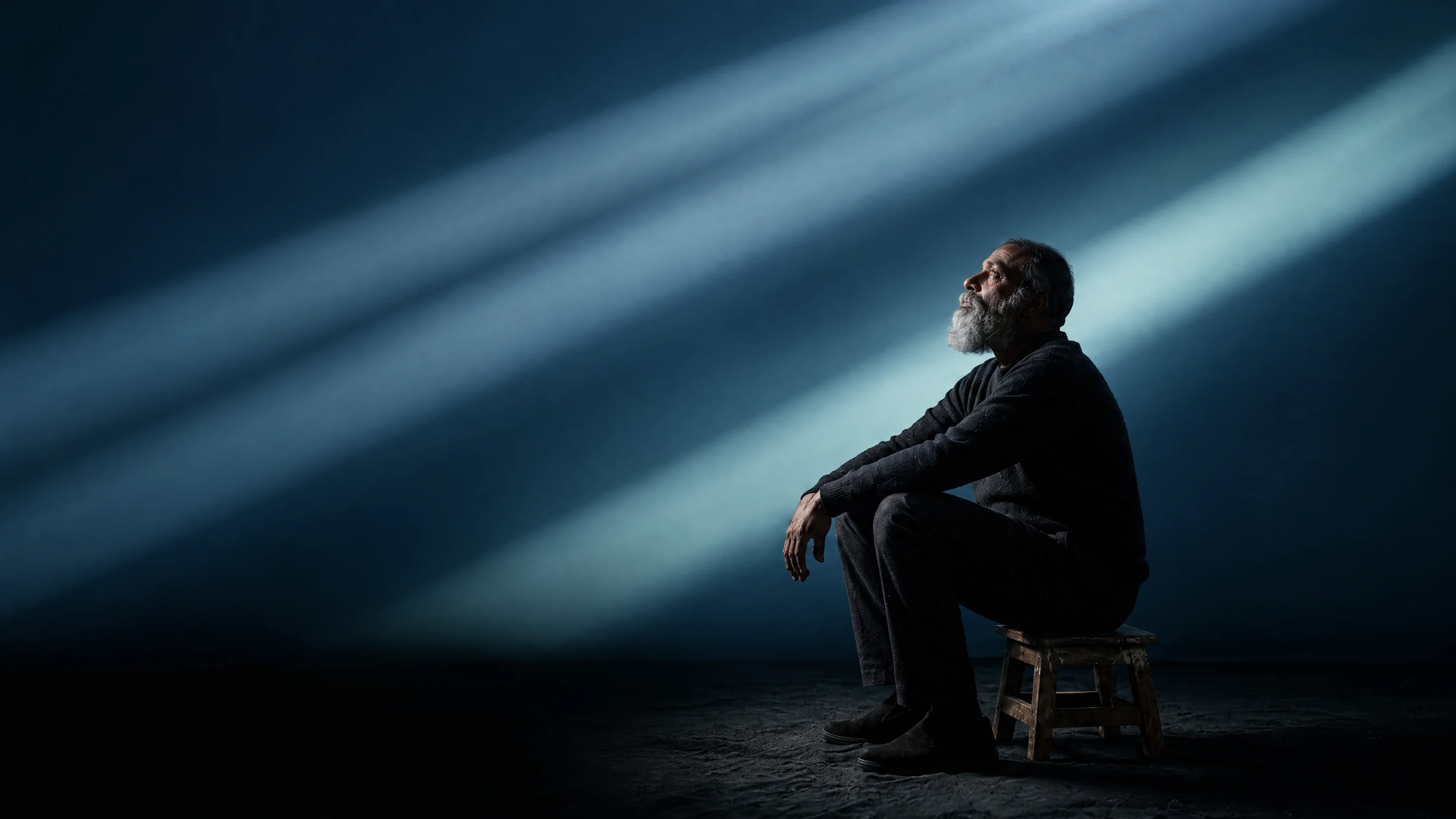 An older man seated on a stool, looking up into beams of blue light