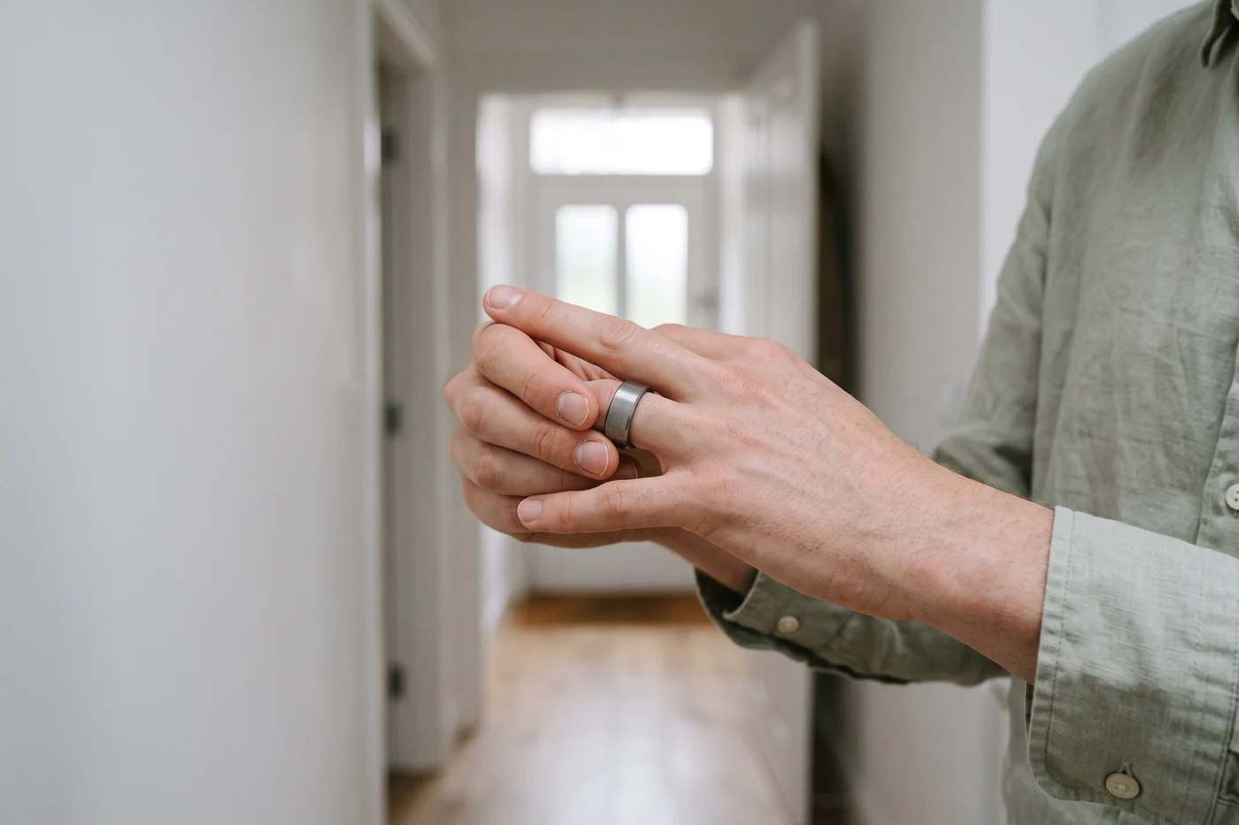 Two hands pushing a wide, flat brushed metal band part way over the knuckle of an index finger, in a hallway with a glazed front door behind.